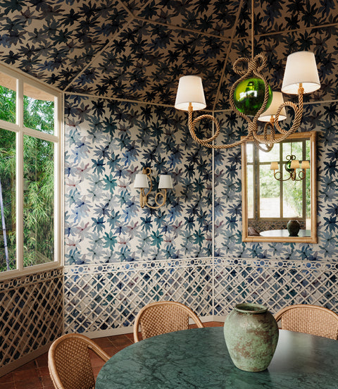 Dining room with blue floral wallpaper covering walls and ceiling, featuring blue leaf patterns; green marble table, wicker chairs, and rustic chandelier with white shades in the foreground.