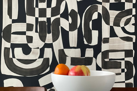 Black and white abstract wallpaper called Down the Rabbit Hole in a dining room setting, with a modern white bowl filled with oranges and an apple placed on a reflective glass table.