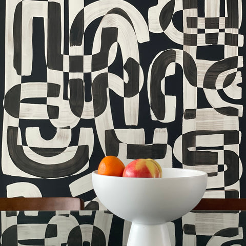Black and white abstract wallpaper called Down the Rabbit Hole in a dining room setting, with a modern white bowl filled with oranges and an apple placed on a reflective glass table.