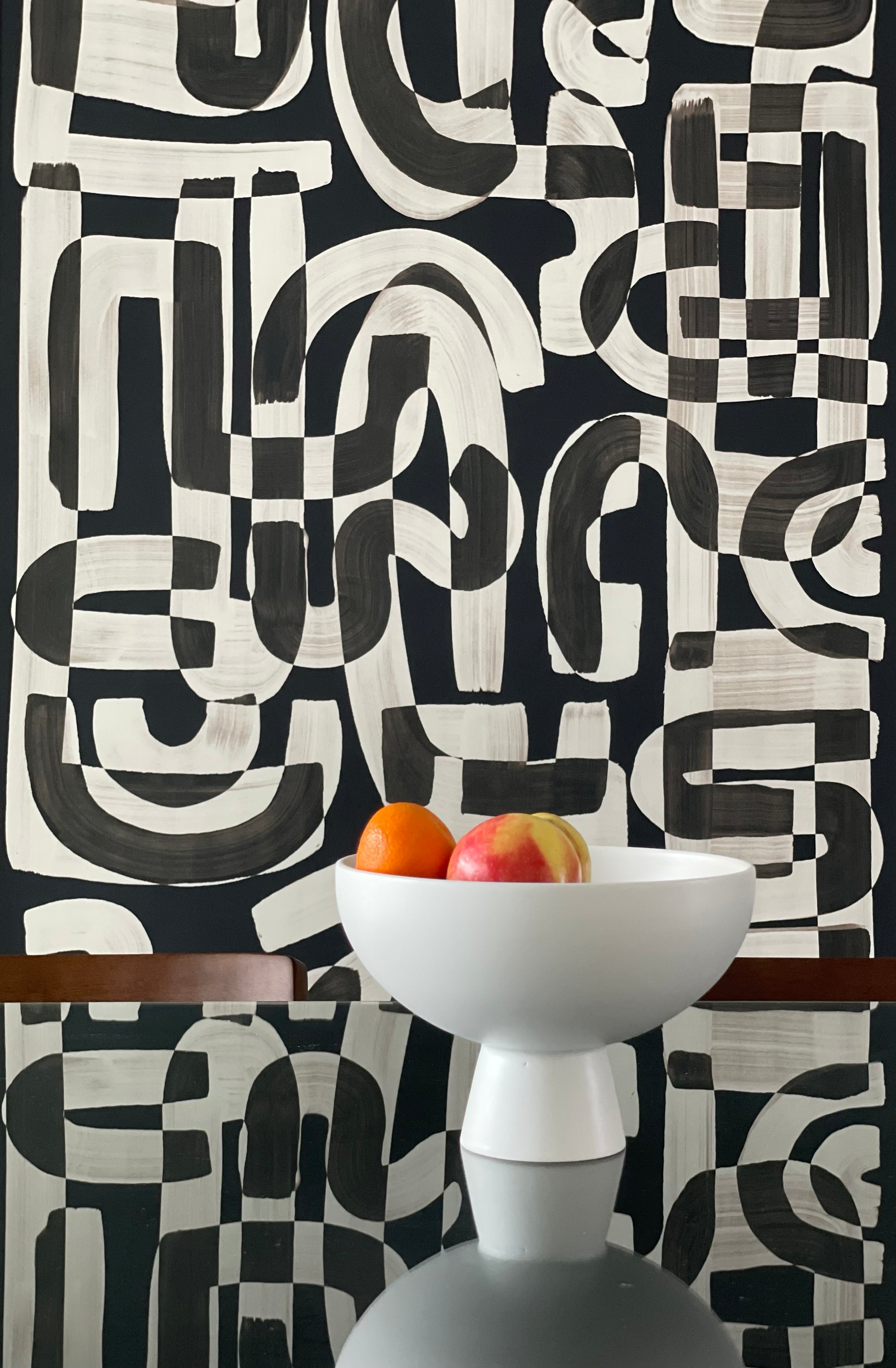 Black and white abstract wallpaper called Down the Rabbit Hole in a dining room setting, with a modern white bowl filled with oranges and an apple placed on a reflective glass table.