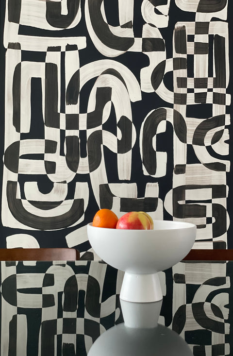 Black and white abstract wallpaper called Down the Rabbit Hole in a dining room setting, with a modern white bowl filled with oranges and an apple placed on a reflective glass table.