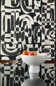 Black and white abstract wallpaper called Down the Rabbit Hole in a dining room setting, with a modern white bowl filled with oranges and an apple placed on a reflective glass table.