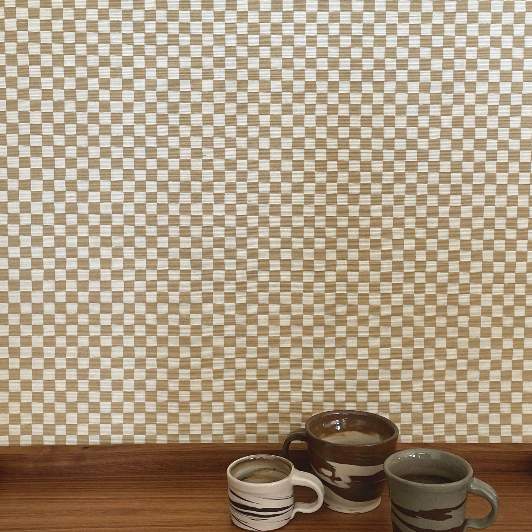 Taupe colored checker grasscloth wallpaper with 3 ceramic mugs on a wooden shelf in bottom of photo