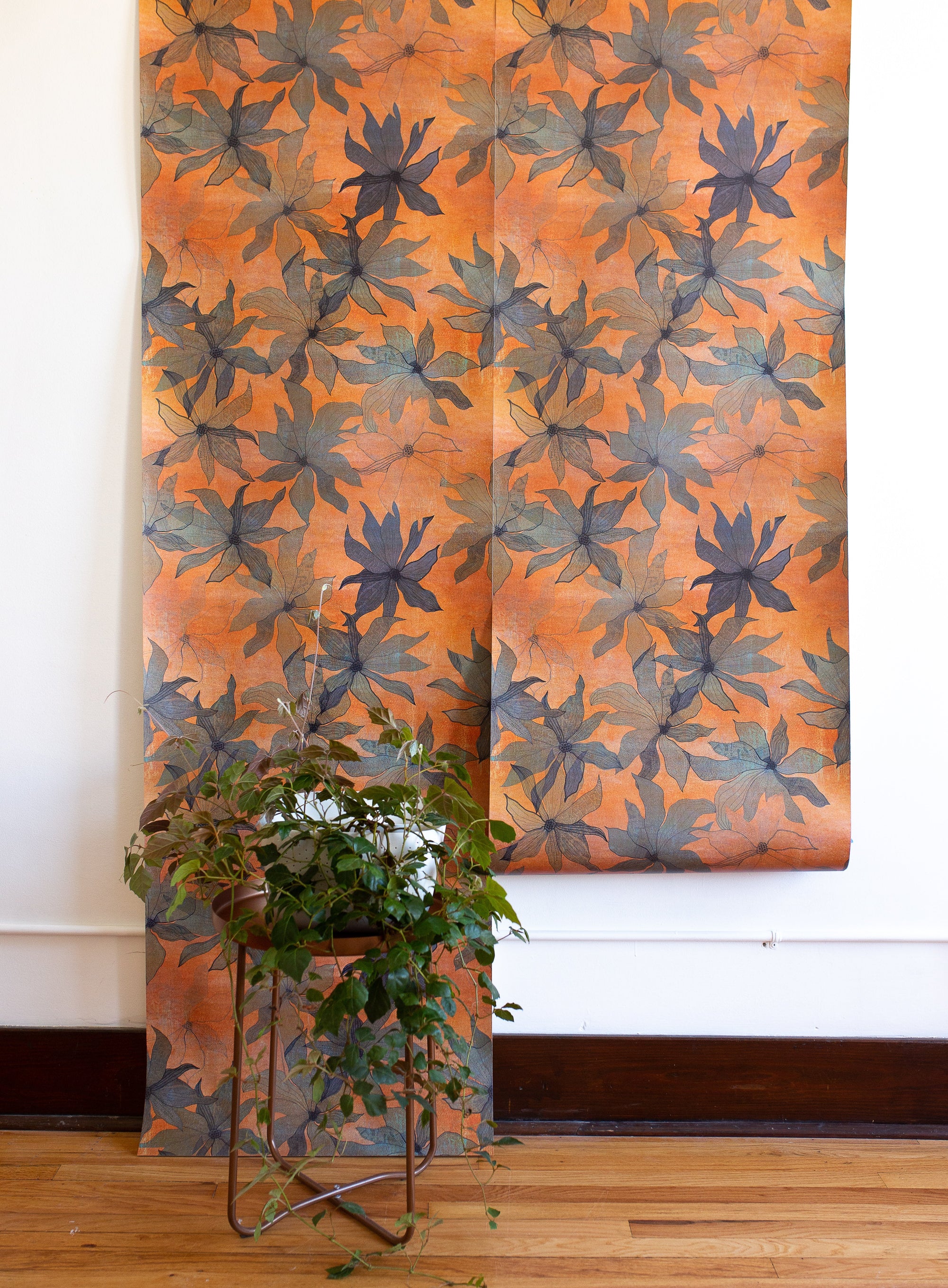 Orange wallpaper with bold floral patterns in muted blue shades, displayed in hanging panels; green leafy plant in a modern metal stand in the foreground.