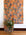 Orange wallpaper with bold floral patterns in muted blue shades, displayed in hanging panels; green leafy plant in a modern metal stand in the foreground.
