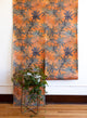 Orange wallpaper with bold floral patterns in muted blue shades, displayed in hanging panels; green leafy plant in a modern metal stand in the foreground.
