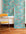 A rolled-out wallpaper featuring a botanical and snake pattern in teal, pink, and gold, displayed against a white wall with a mustard yellow vintage chair.