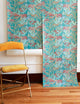 A rolled-out wallpaper featuring a botanical and snake pattern in teal, pink, and gold, displayed against a white wall with a mustard yellow vintage chair.